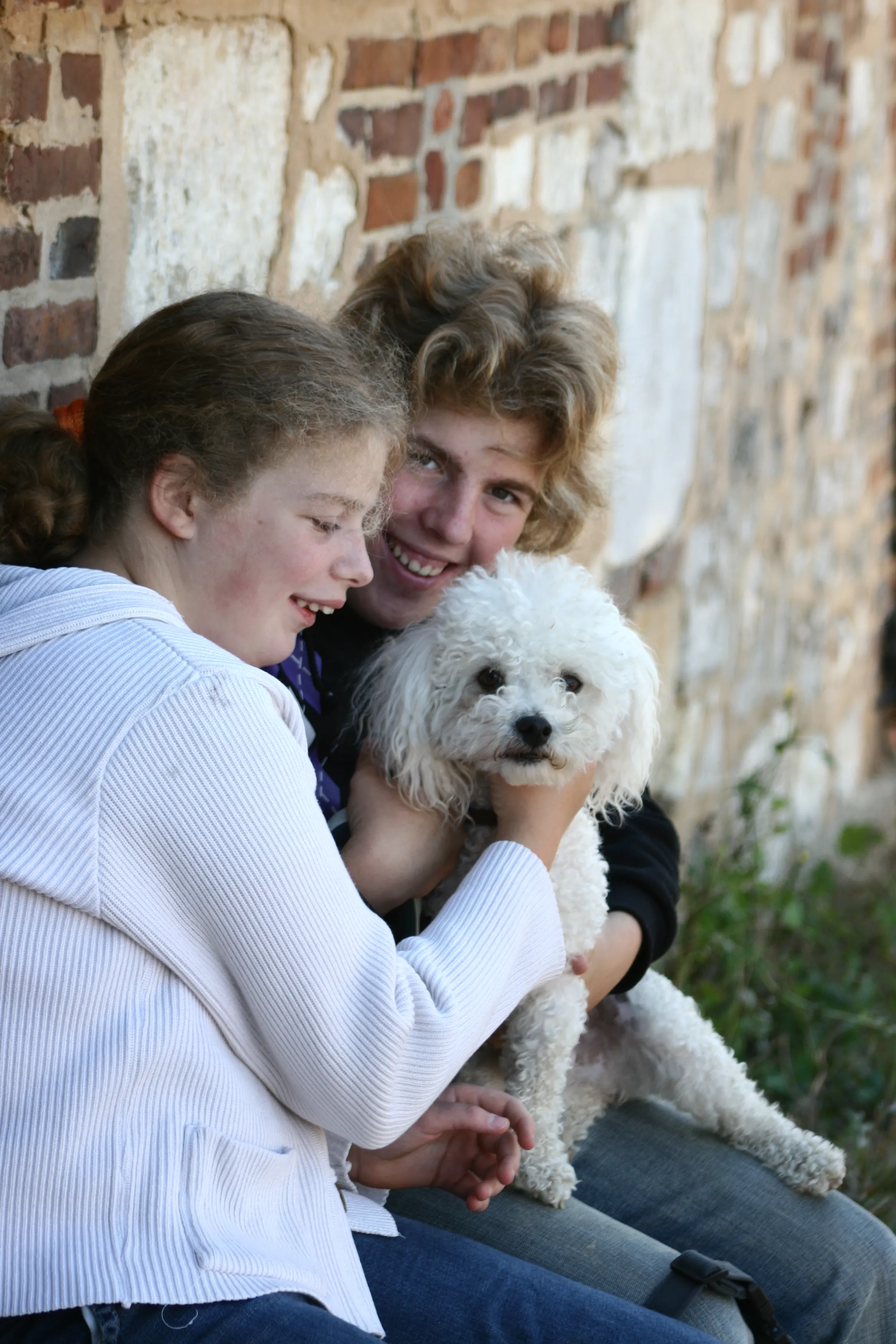 Two siblings sitting against a brick wall holding a small white poodle